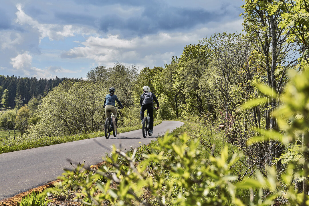 Zwei Radfahrer fahren genüsslich auf dem mit Feinsphalt belegten Vulkanradweg im hessichen Vogelsberg. Die Randstreifen sind üppig grün und die Laubbäume beschatten die sonnige Trasse. Man kann noch erkennen, dass der Radweg auf dem ehemaligen Bahndamm der Oberwaldbahn liegt, die hier einst in den 70er Jahren von Ort zu Ort fuhr.