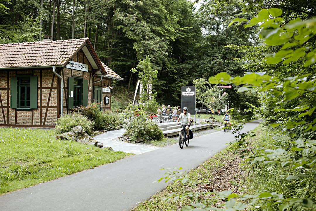 Radfahrer fahren auf dem geteerten Vulkanradweg an der Zentralstation in Lauterbach-Frischborn vorbei, die mitten in einem Waldstück liegt. Die Zentralstation ist der ehemalige Bahnhof und wird heute gastronomisch mit einem Biergarten genutzt.