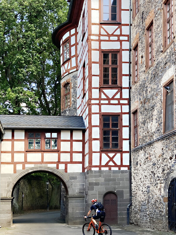 Ein sportlicher Radfahrer fährt auf den Torbogen des Fachwerkanbaus an Schloss Laubach zu. Das zu überwiegenden Teilen aus Basaltstein errichtete Schloss befindet sich inmitten der Fachwerkaltstadt von Laubach und durch den Torbogen gelangt man hinter das Schloss, wo sich ein weitläufiger Park erstreckt.