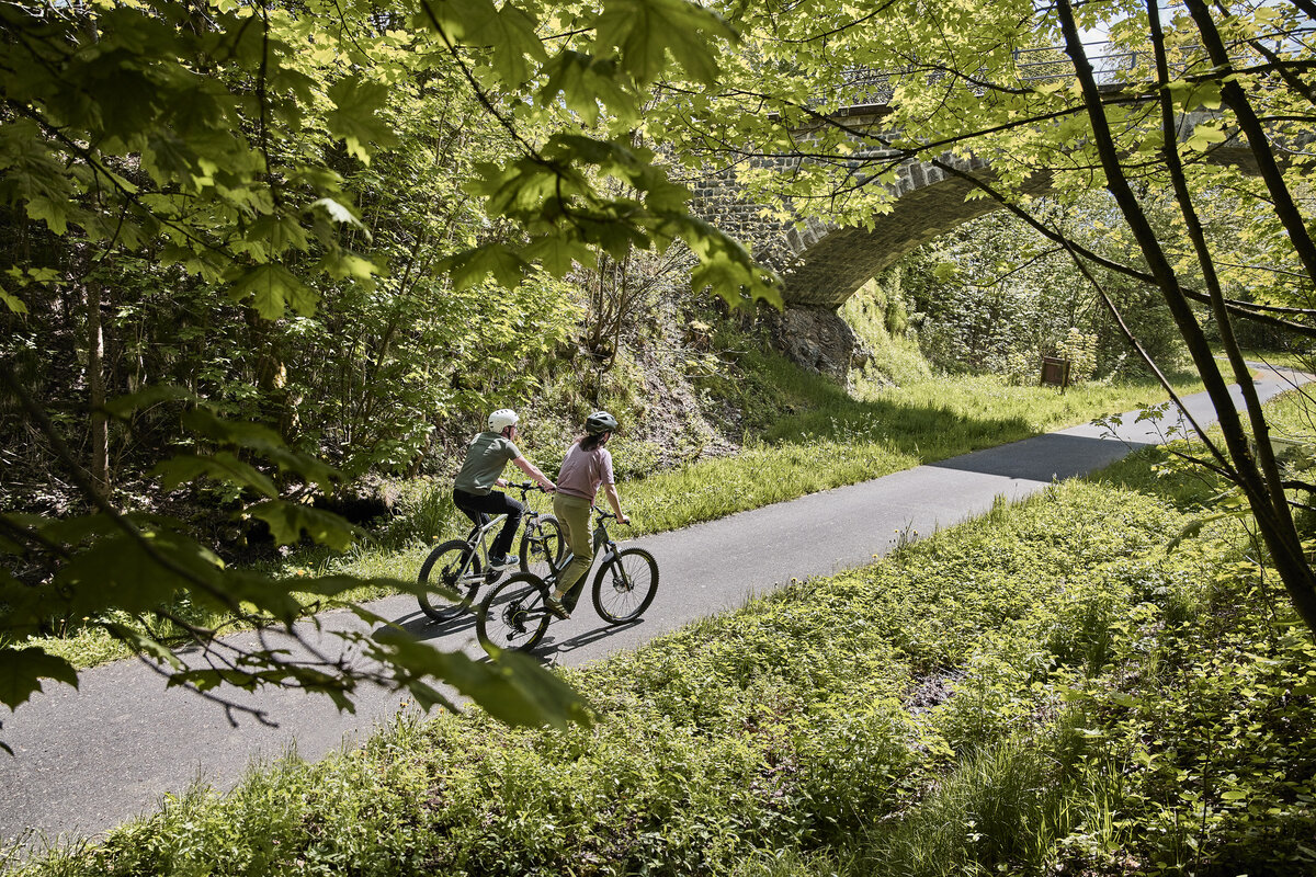 Zwei Radfahrer fahren genüsslich auf dem mit Feinsphalt belegten Vulkanradweg im hessichen Vogelsberg. Die Randstreifen sind üppig grün und die Laubbäume beschatten die sonnige Trasse. Vor den Radlern liegt eine Basaltbrücke unter der der Radweg hindurchführt.