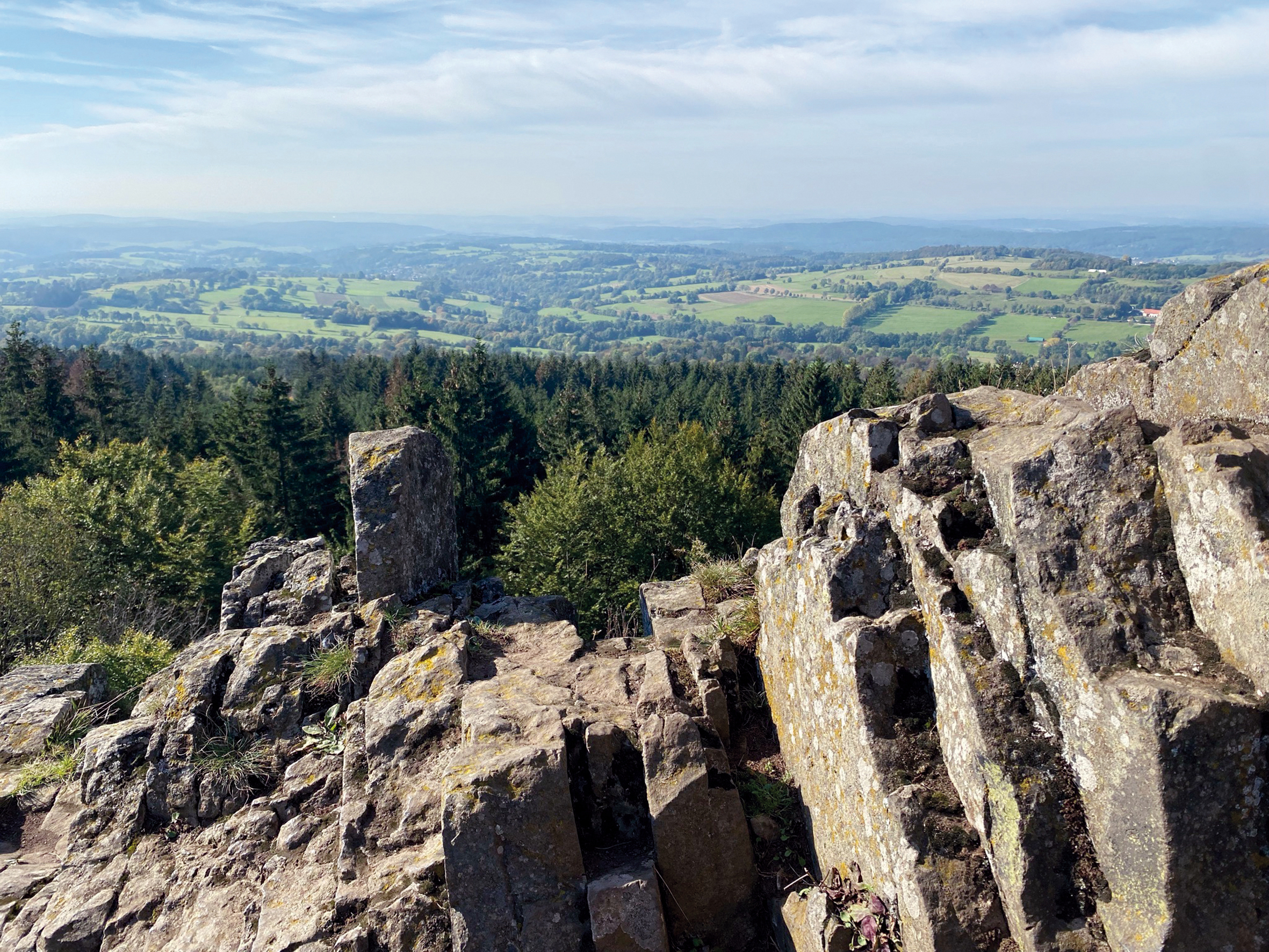 Der Blick vom schroffen Basaltfelsen des Geotopes Bilstein im hessischen Vogelsberg geht weit über das geschwungene Land der Vulkanregion. Die sommerlich grünen Felder, Wiesen und Wälder stehen im Kontrast zum braun-grauen Basaltgestein des Berges Bilstein.
