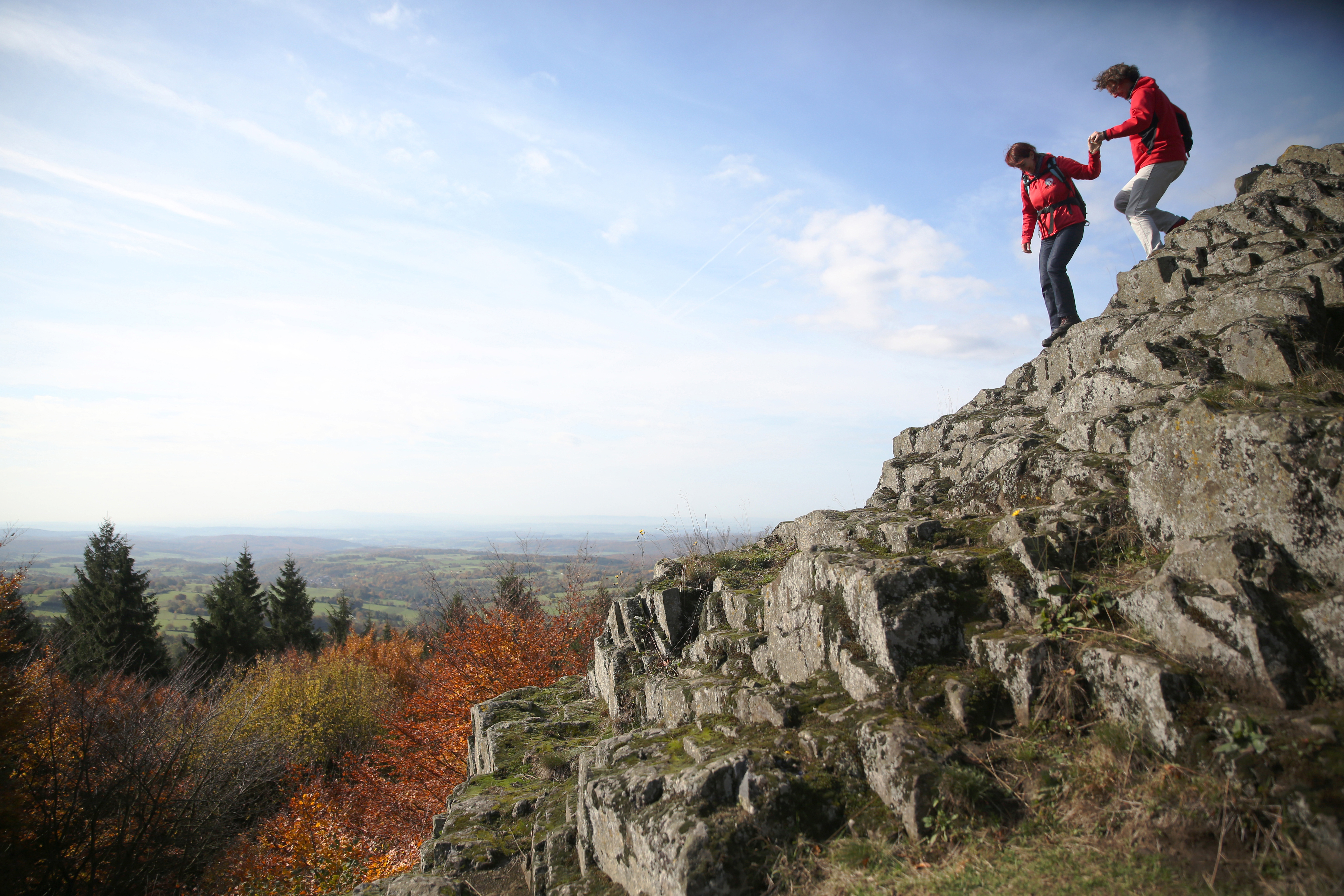 Auf den schroffen Basaltfelsen des Geotopes Bilstein im hessischen Vogelsberg steigen zwei Wanderer Hand in Hand den Berg hinab. Vom Berg aus geht der Blick weit über das geschwungene Land der Vulkanregion. Der Herbst hat das Laub des Waldes bunt gefärbt.